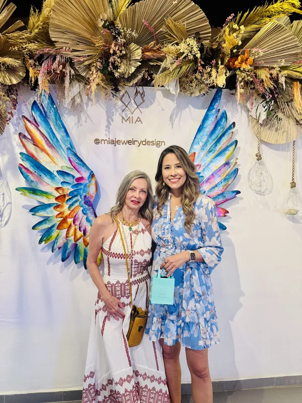 Two guests in white and blue dresses holding an aqua MIA gift bag, framed by the painted angel wings at a MIA event