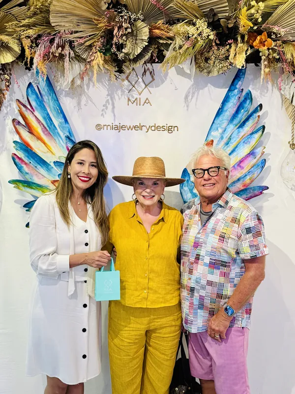 Imelda in white with two collectors under a dried-flower arch; the client in yellow holds her aqua MIA gift bag
